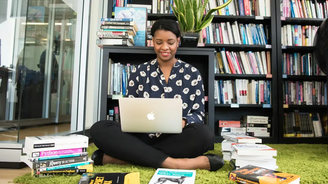 A person sits cross-legged on a green rug in a library with a laptop on their lap, surrounded by stacked books.