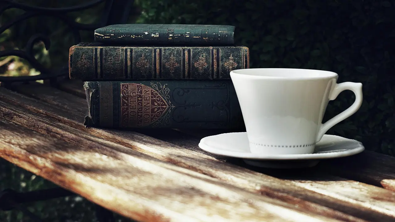Stack of vintage hardcover books on a weathered wooden table with a white teacup and saucer, outdoors.