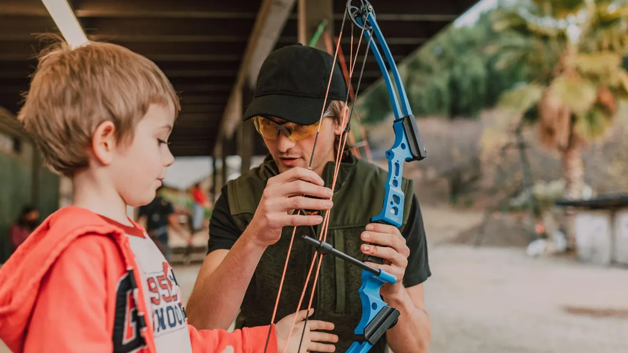 A young boy in a red hoodie watches a man wearing a black cap handle a blue toy bow with strings under a sheltered outdoor area.