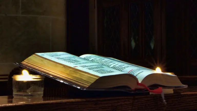 An open book resting on a dark wooden surface, with small lit candles on either side, bathed in warm glow.
