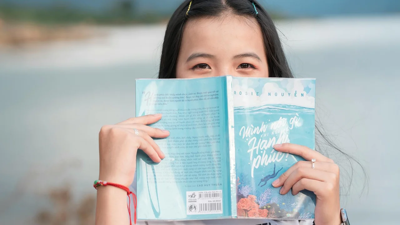Young woman at the beach holding a blue book in front of her face, eyes visible as she reads.
