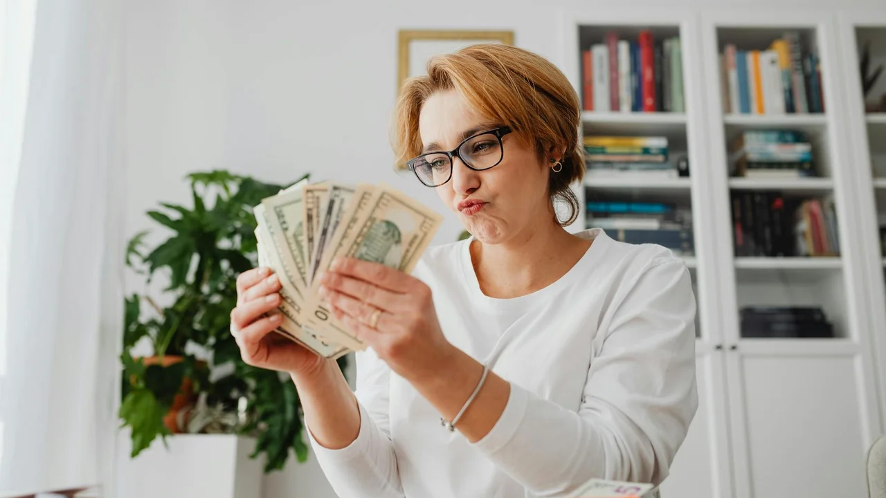 Woman with glasses counts money in a bright room with a bookshelf in the background.