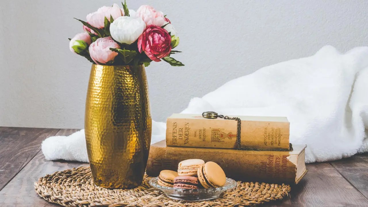 A golden vase with pink roses sits on a wooden table beside a stack of vintage books tied with string and a plate of macarons, with a white cloth in the background.